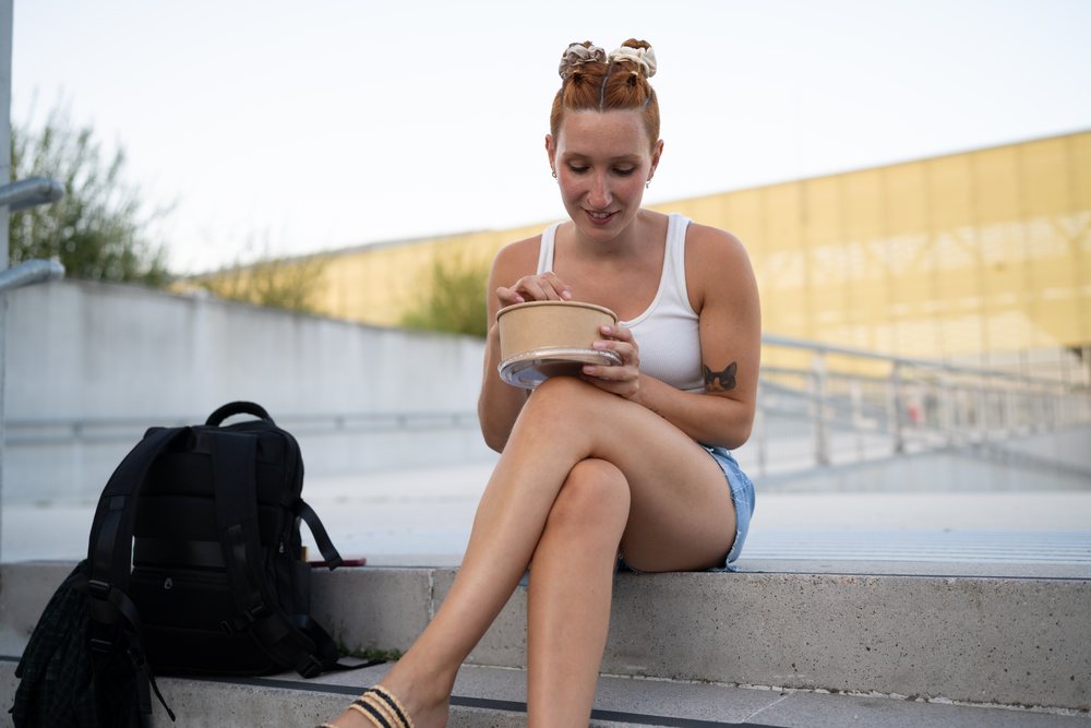 A young woman with red hair is eating from a food container while sitting on concrete steps in an urban environment, Santiago De Compostela, Spain.