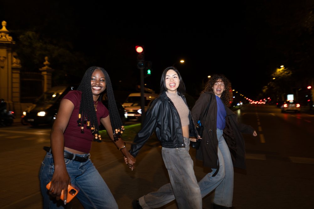Three women joyfully holding hands while walking across a city street at night under a red traffic light, Madrid, Spain.
