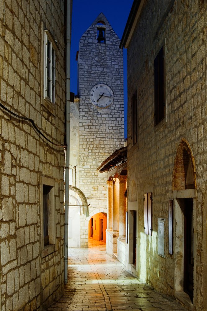 Stone clock tower and illuminated narrow cobblestone alleyway with shuttered stone buildings and an archway at night, Rab Island, Croatia.