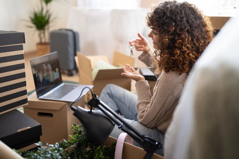 Woman with curly hair sitting on the floor holding keys during a video call amid unpacked moving boxes in a home interior, Spain.