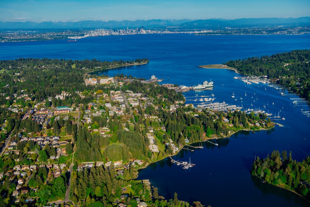 Aerial view showing Bainbridge Island town, its marina with boats, and city skyline across Puget Sound under a clear blue sky, Salish Sea, Eagle Harbor, USA.