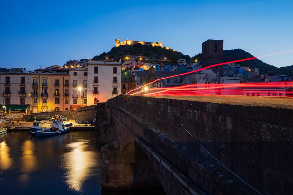 The stone bridge spans the Temo River at Dawn, with boats moored near the riverbank and the illuminated hilltop castle overlooking, Travel Images, Bosa, Sardinia, Italy.