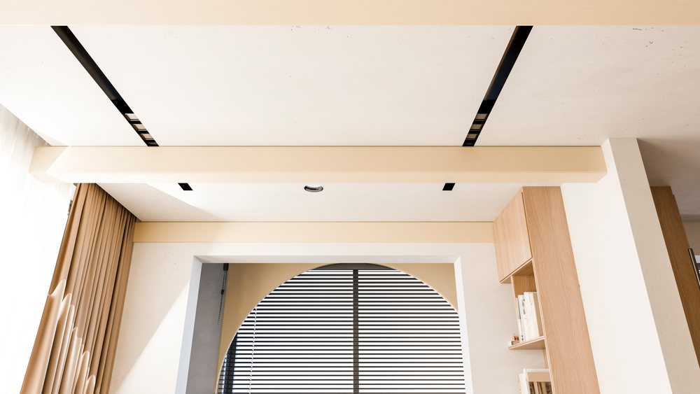 Modern interior featuring ceiling beams, an arched window with blinds and beige curtains, and a wooden bookshelf with books.