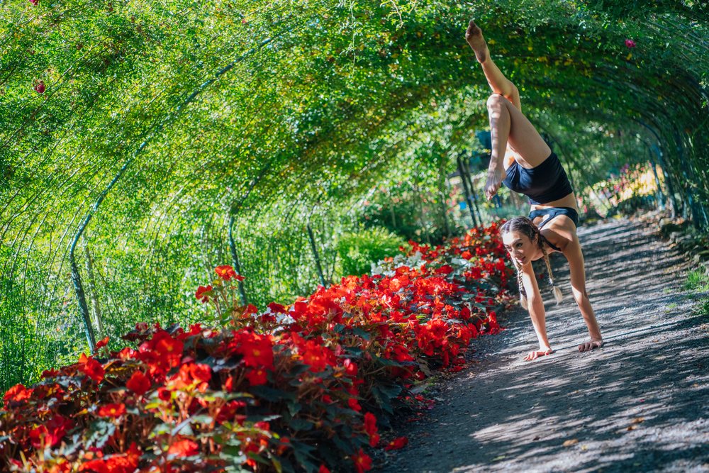 Woman contortionist performs a handstand on a dirt walking path beside red flowers under an arched trellis in a green garden, Tacoma, WA, USA.