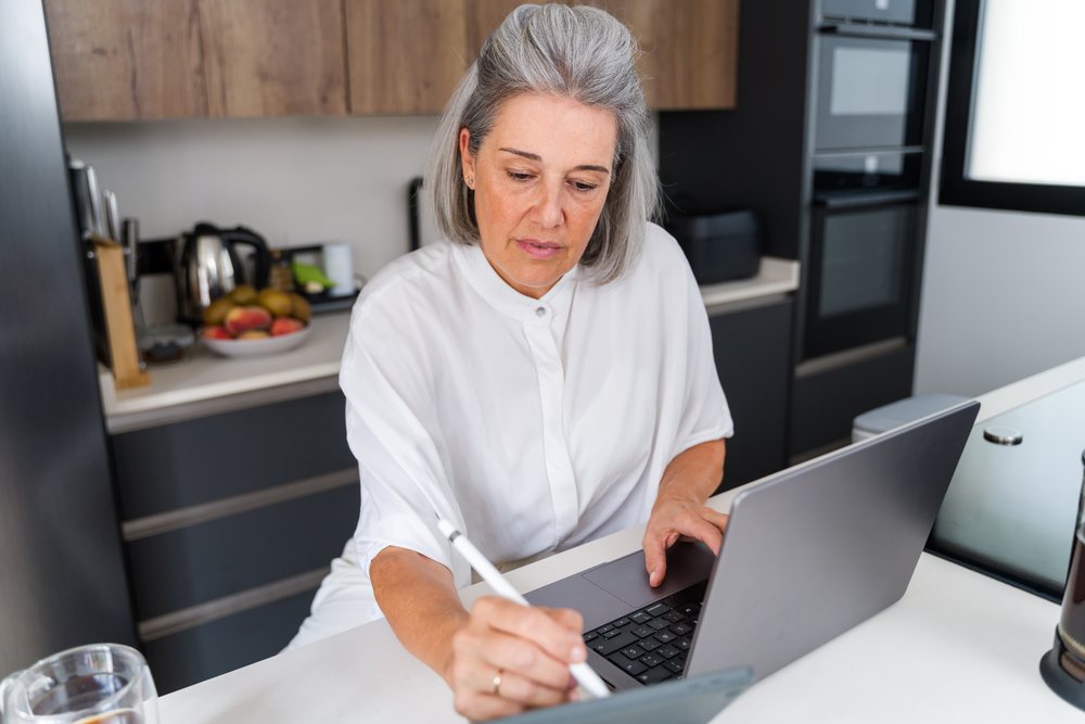 Woman using a laptop and writing on a tablet with a digital pen in a modern kitchen, Spain.