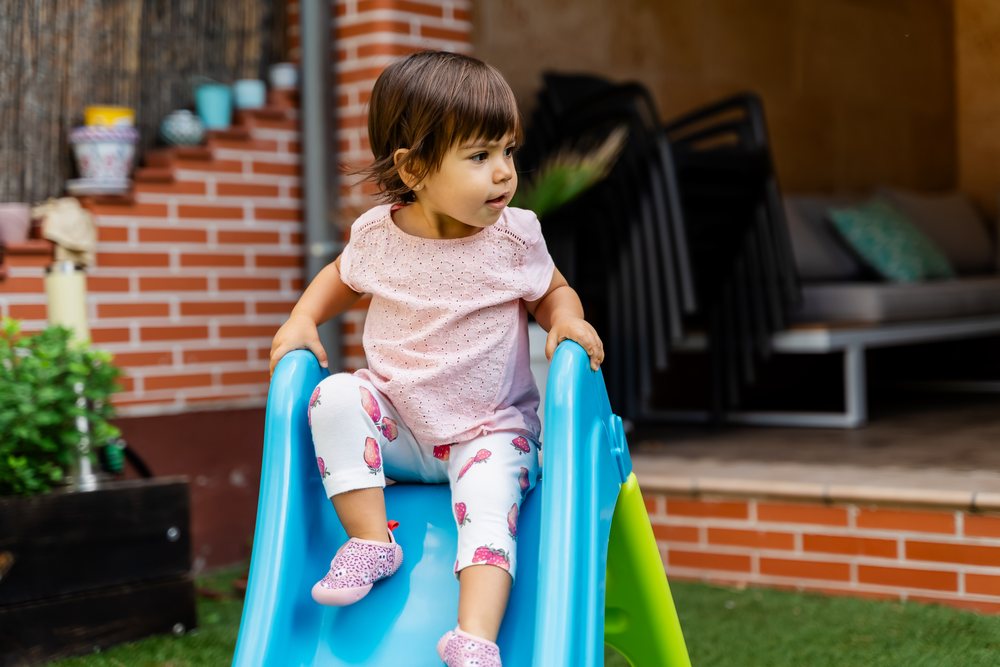 Toddler girl wearing strawberry leggings and pink shirt sitting on a blue plastic slide in a backyard playground, kids, Spain.