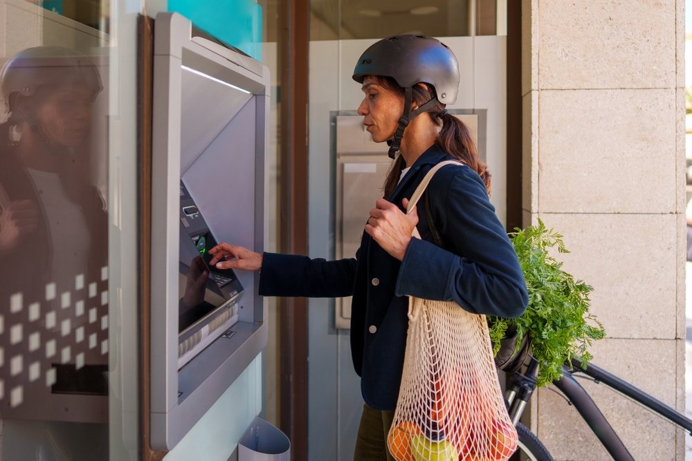 Woman wearing a helmet uses an outdoor ATM beside her bike while carrying a net bag with groceries, Málaga.