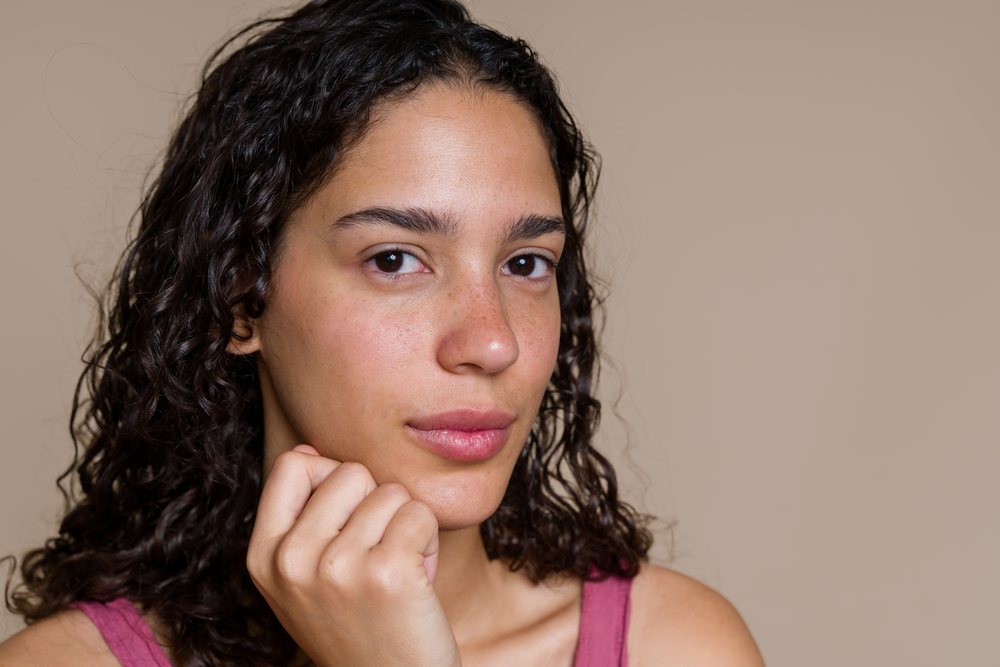 Young adult woman with curly hair, freckles, and natural skin poses for a beauty shoot in a studio with light brown background, Spain.