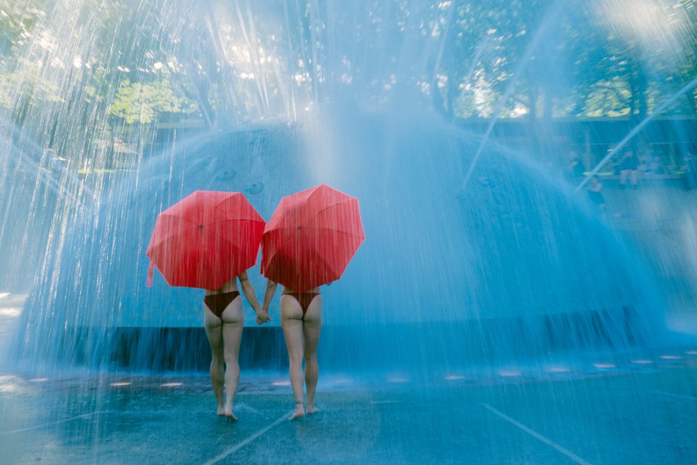 Two women holding hands and carrying umbrellas walk barefoot through a water fountain spray in a public park, partner, acrobatics, yoga, USA.