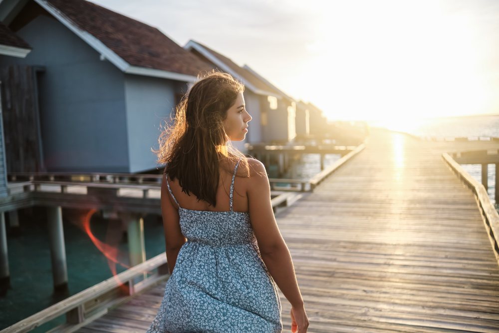 A woman in a blue summer dress walks along a wooden boardwalk past overwater bungalows at sunset in the, Maldives.