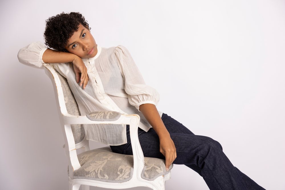 Woman with curly hair wearing a white blouse and dark jeans sits relaxed on a white decorative chair in a studio portrait, fashion, young culture.