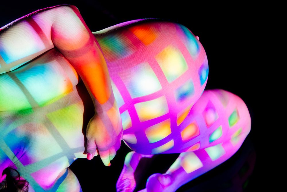 Pregnant woman kneels nude in a black studio, her body decorated with a colorful multicolor grid light projection, pregnancy.