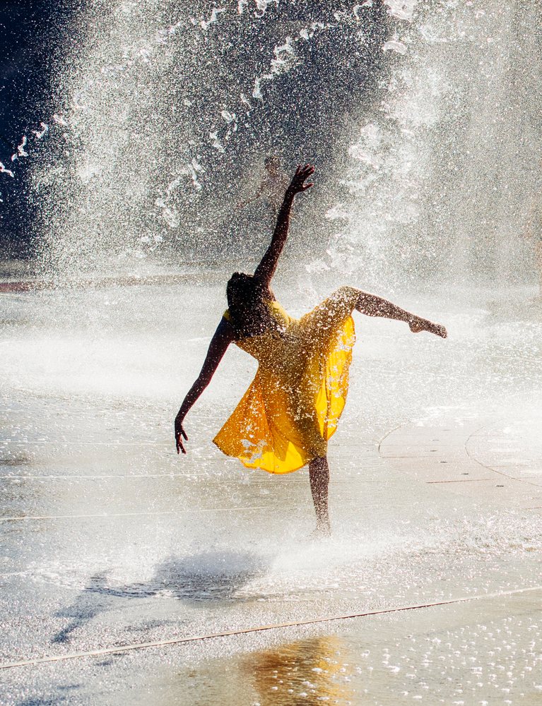Female dancer in a yellow flowing dress performs a graceful pose amid water spray from an outdoor fountain, USA.