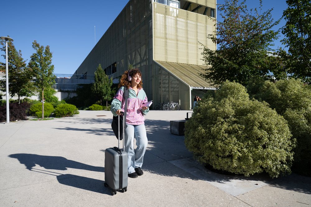 Woman walking outdoors pulling luggage and holding a notebook near a modern building with trees, Spain.