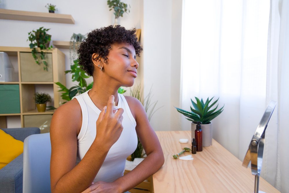 Young woman sprays perfume at a wooden vanity table with skincare tools and decorative plants in a bright indoor home setting.