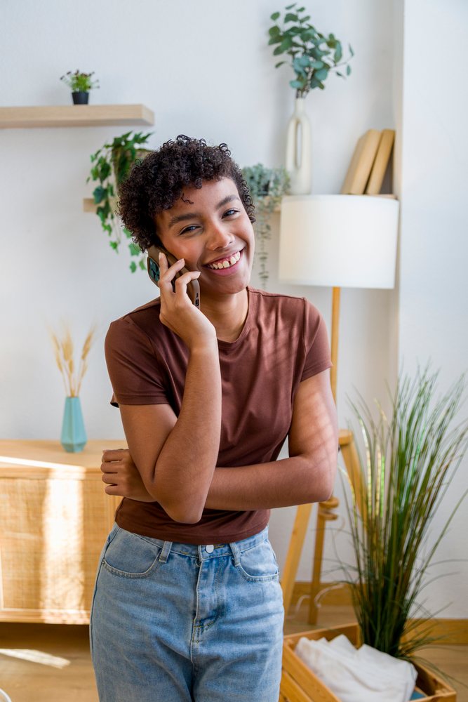 Young woman with curly hair smiling and talking on a mobile phone in a modern, well-lit home interior with plants and wooden furniture.