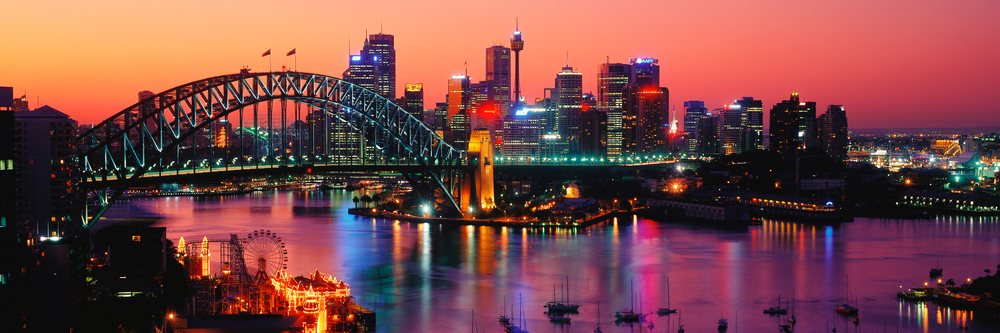 Sydney skyline at dawn featuring illuminated Harbor Bridge, boats in the harbor, and a lit ferris wheel reflecting on the water in Australia.