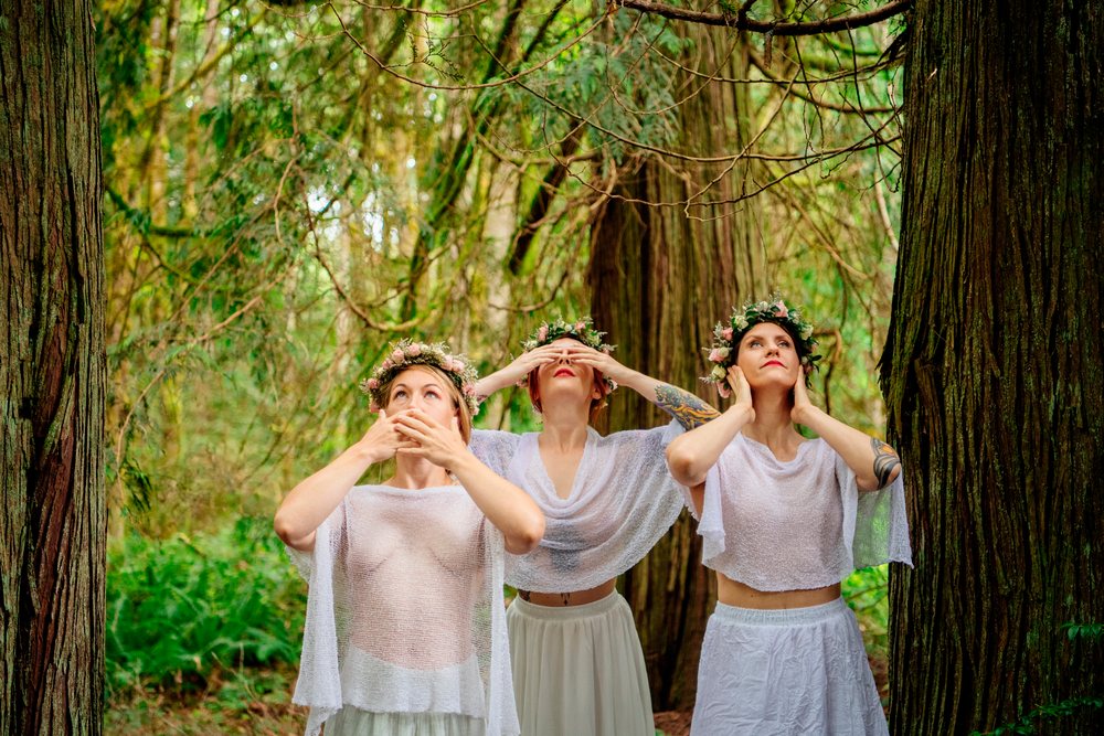 Three women in white sheer tops and skirts wearing floral crowns stand in a wooded forest surrounded by tall trees and greenery, muses, USA.
