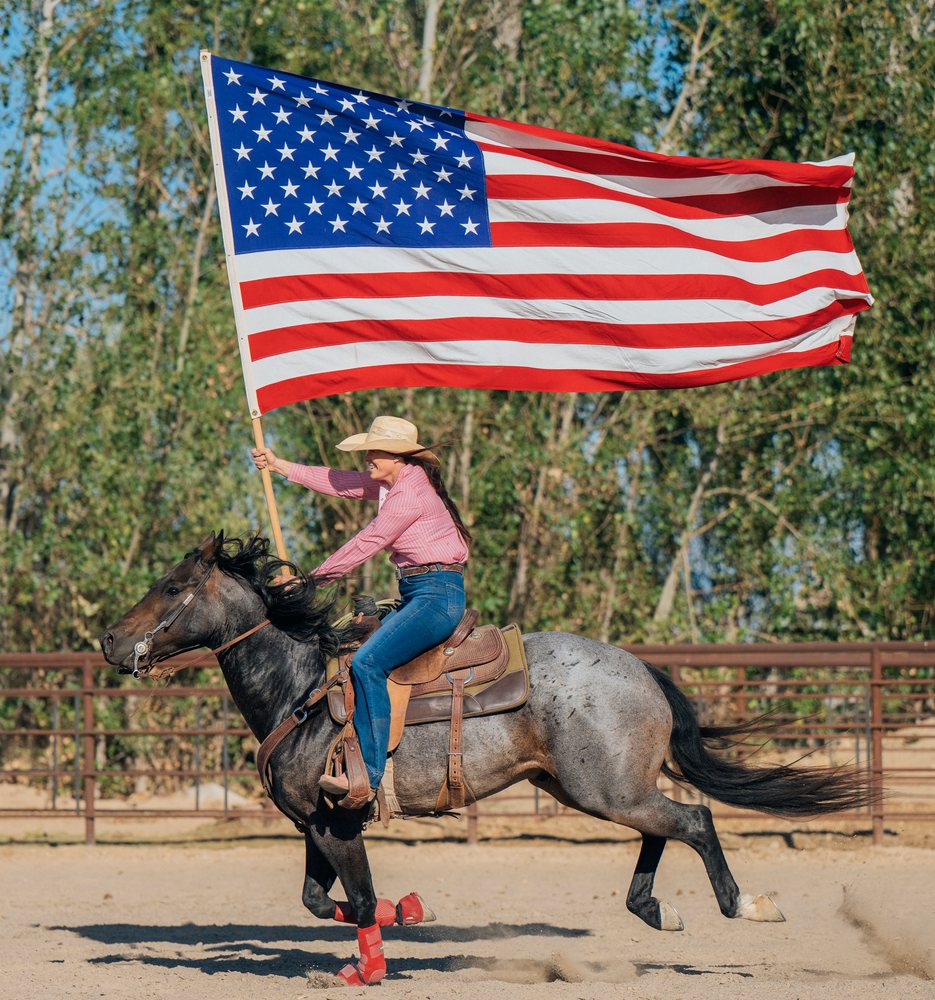 Woman wearing cowboy hat and jeans rides galloping horse holding large American flag in outdoor rodeo arena with green trees, Southwest USA.
