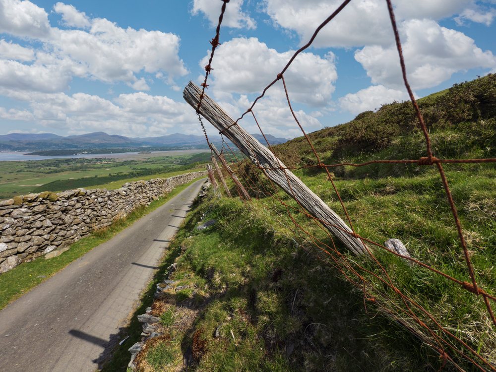 Rural narrow paved road beside wire fence and stone wall along grassy hillside overlooking scenic Welsh countryside, Landscape, Harlech, Gwynedd, Wales.