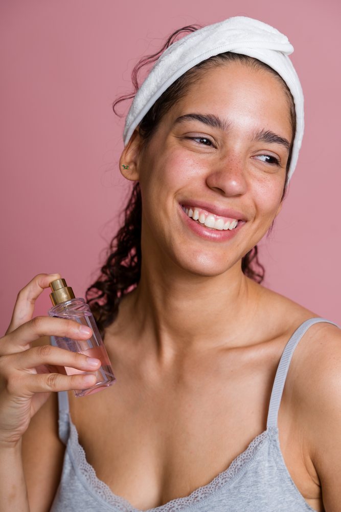 Woman smiling and holding a clear facial spray bottle in a beauty shoot with a pink background, Spain.