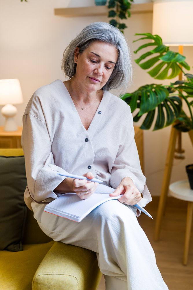 Middle-aged woman with grey hair writes in a notebook while seated on a yellow couch in a modern indoor living room with a monstera plant and lamp, Spain.
