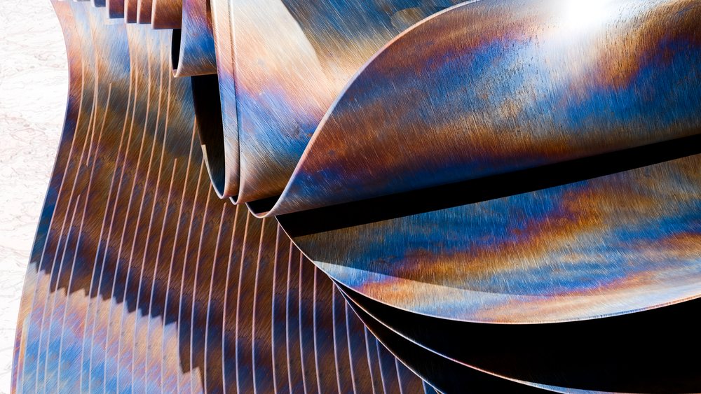 Folded rusted metal sheets with blue and orange oxidation patterns create an abstract sculptural texture in an industrial outdoor setting.