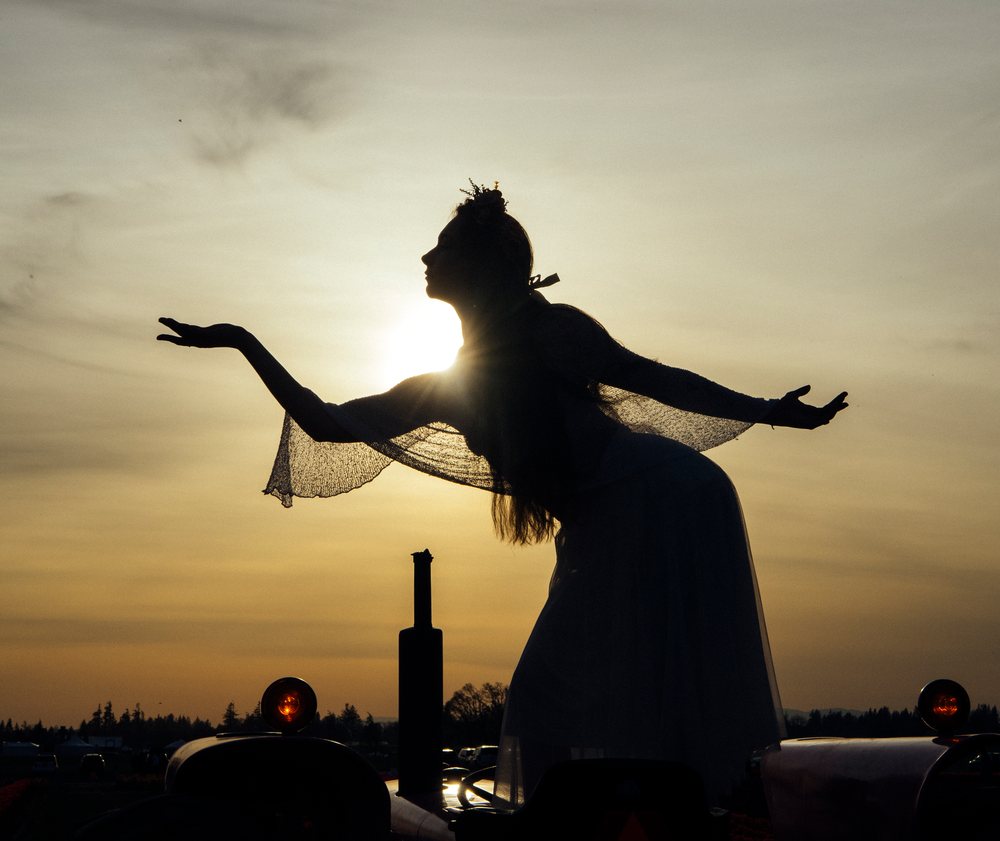 Silhouette of a woman wearing a crown poses outdoors at sunset in a natural USA setting with trees and a vehicle visible.