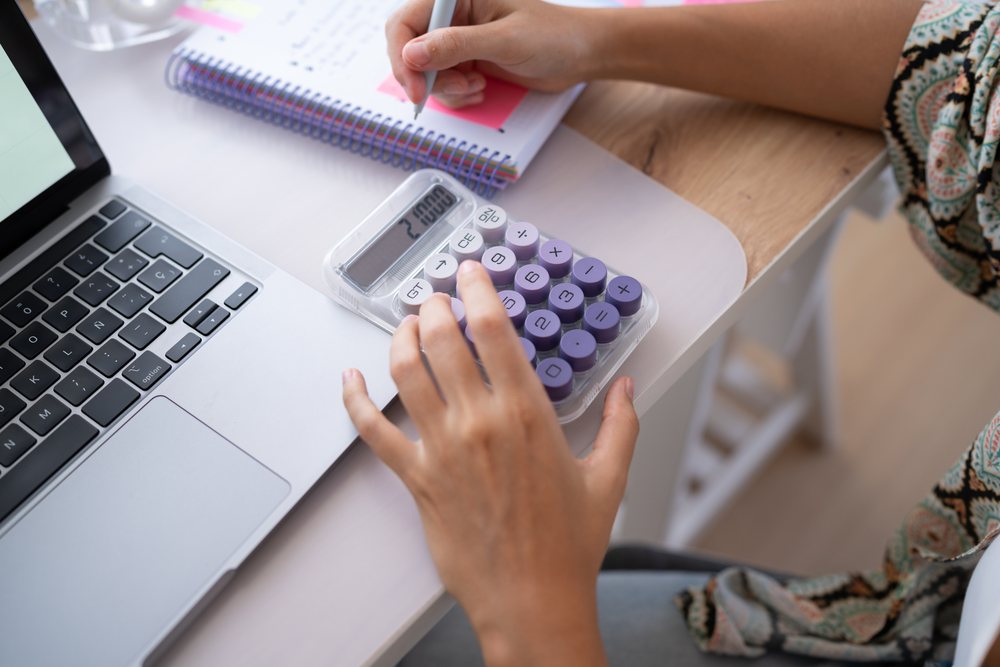 Hands operating a calculator and writing notes on a spiral notebook beside a laptop on a desk in a home office, Spain.