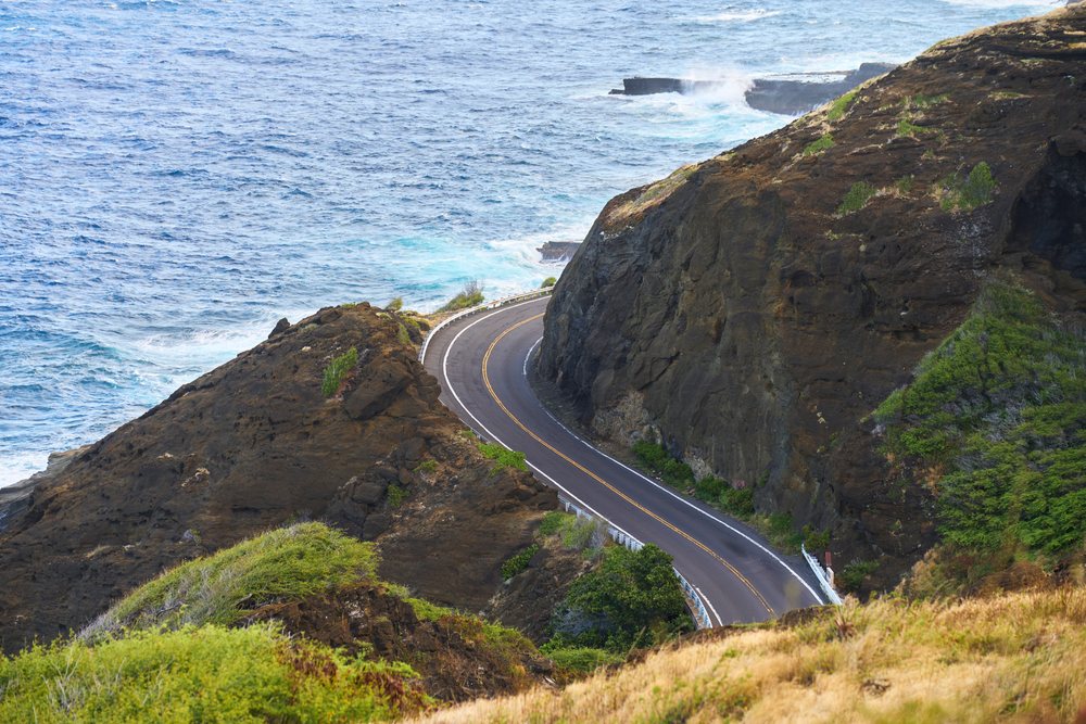 Coastal road curves between cliffs above the ocean, with waves crashing against the rocks, Oahu, Hawaii, USA.