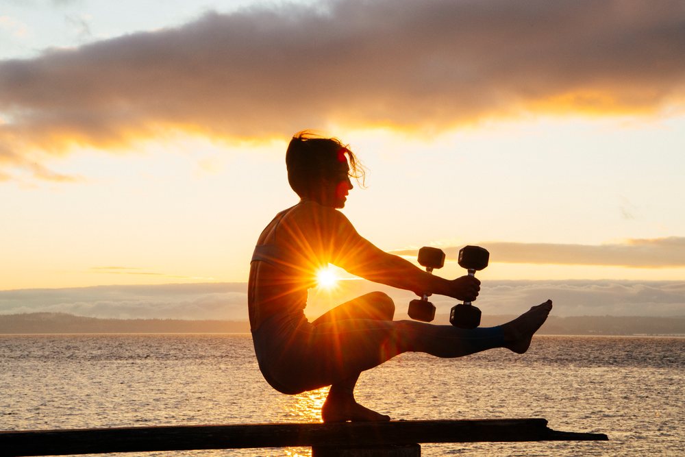 Person performing a yoga balance pose holding dumbbells barefoot at sunset by calm water, WA, USA.