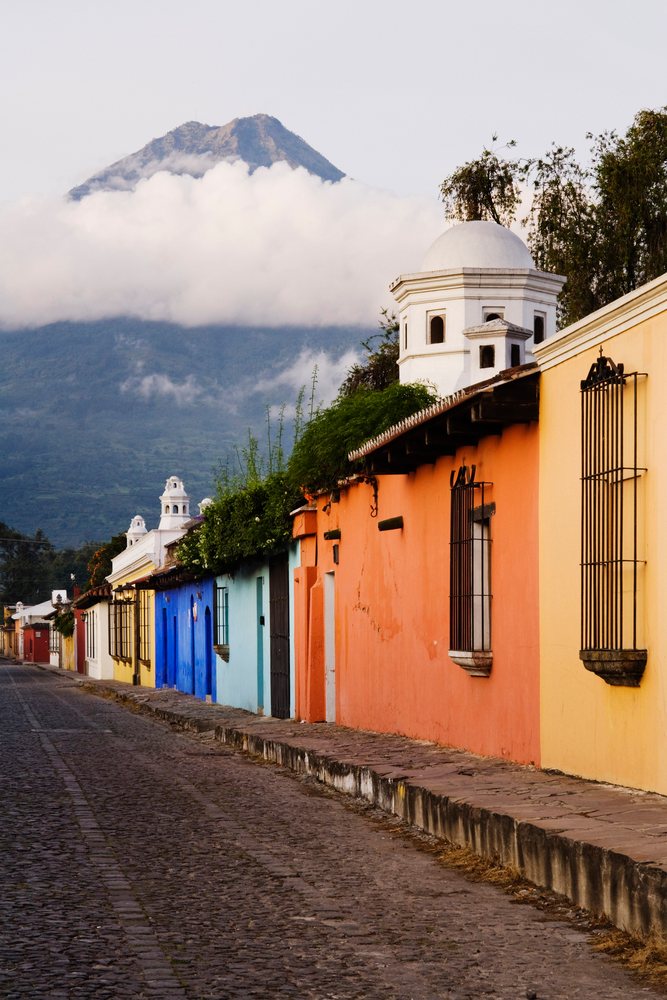Agua Volcano rises behind colorful colonial buildings and a cobblestone street in historic, Antigua, Guatemala.
