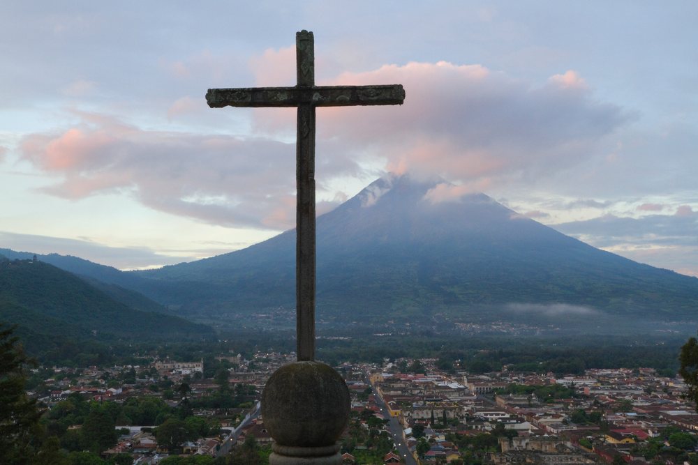Cross on stone pedestal at Cerro de la Cruz overlooking Antigua town and Volcan de Agua volcano at dawn with cloudy sky, Sacatepequez Department, Guatemala.
