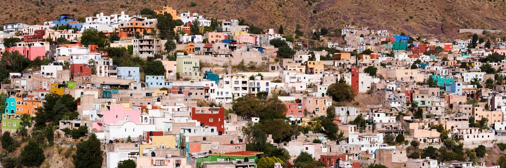 Multistory colorful houses densely built on a steep hillside slope in Mexico with dry mountainous terrain and visible natural vegetation.