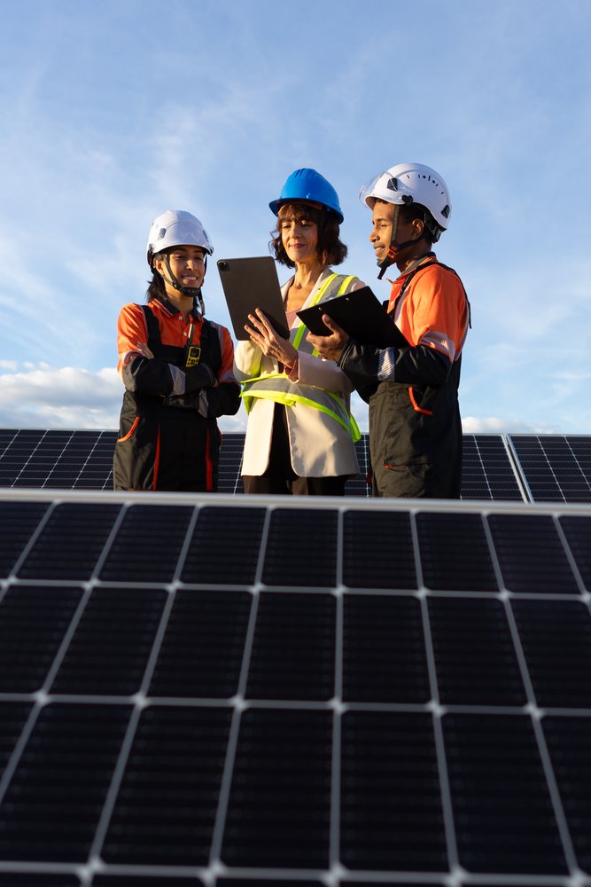 Three workers wearing hard hats and safety vests discuss solar panels outdoors under a blue sky, Madrid, Spain.