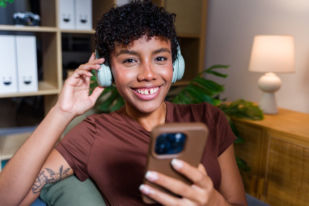Woman with short curly hair wearing headphones and holding smartphone, smiling indoors on a couch near a lamp and plant, Spain.