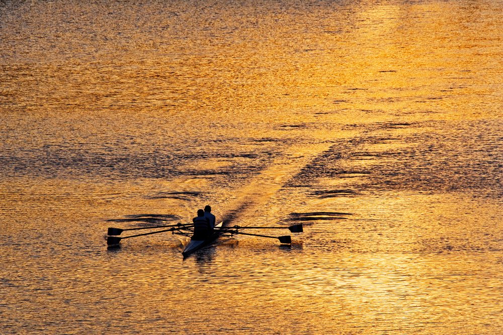 Two people rowing a narrow boat on calm water with golden light reflections, Italy.