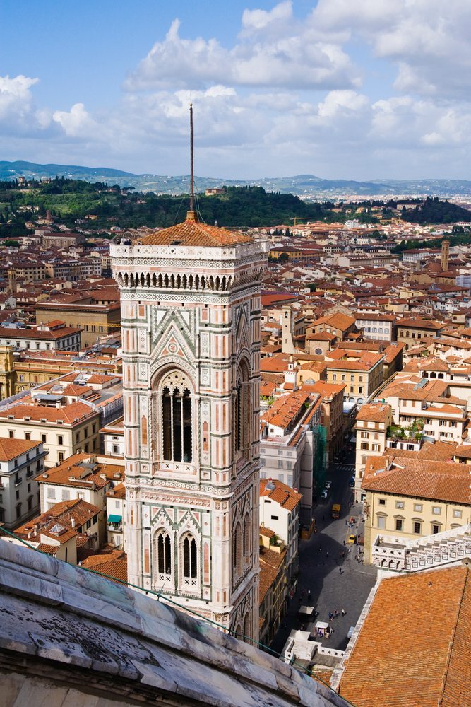 Campanile Cathedral tower rises above Florence with historic red-tiled roofs and hills in the background on a clear day, Italy.