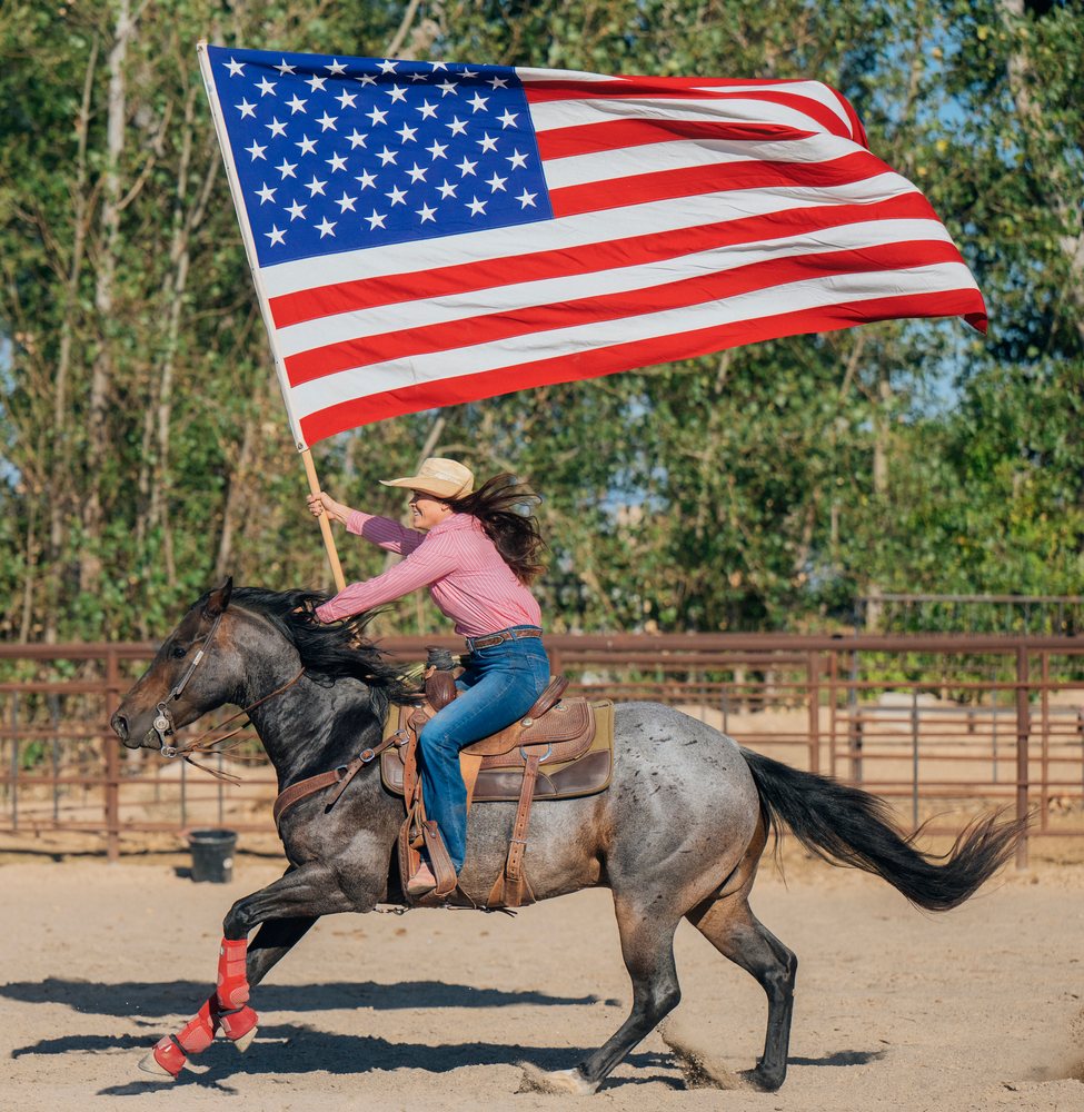 Woman wearing a straw cowboy hat gallops a dapple gray horse while holding a large waving American flag in an outdoor arena, Southwest USA.