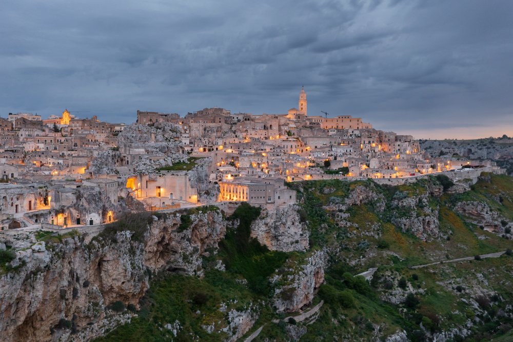 Illuminated historic stone buildings atop rocky cliffs in Sasso Caveoso at Dawn under a cloudy sky with greenery on the cliffs, Matera, Basilicata, Italy.
