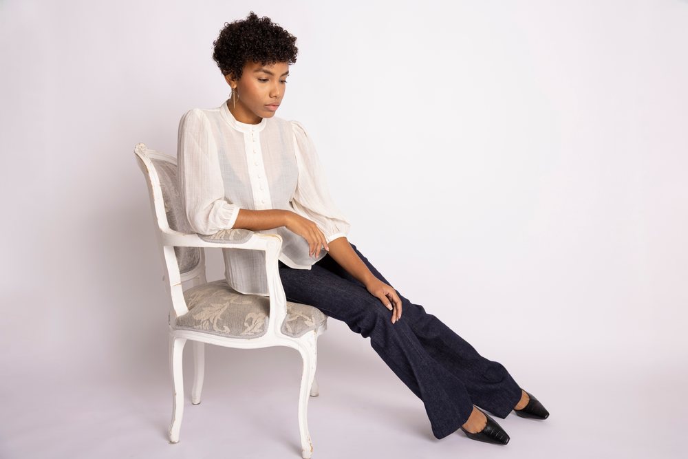 Young woman with curly hair wearing a white blouse and dark jeans seated on a vintage chair in a studio fashion portrait.
