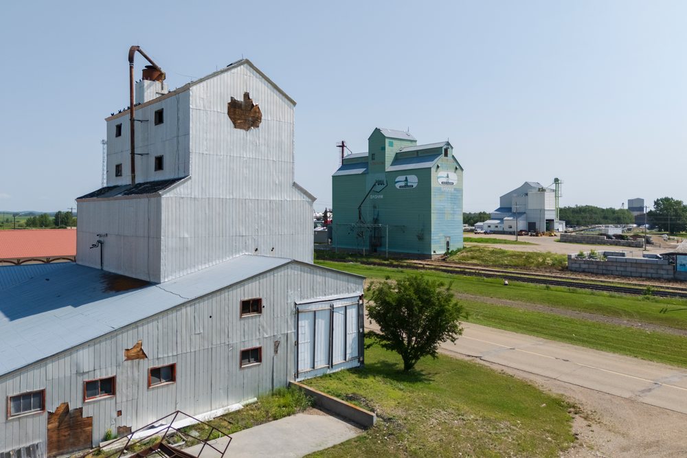 Metal-sided grain elevator and industrial buildings stand beside railroad tracks and a road in rural Canada under a clear summer sky, Storm Chasing, Weather, Environment, USA.
