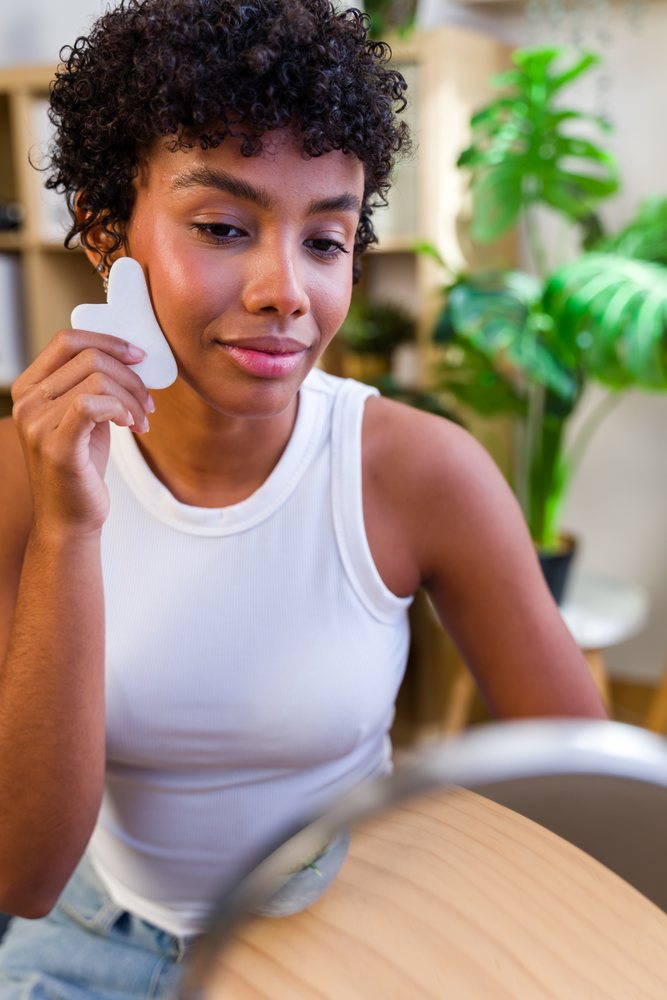 Woman with curly hair wearing a white tank top uses a gua sha tool for facial massage at home beside a mirror and an indoor plant, Spain.