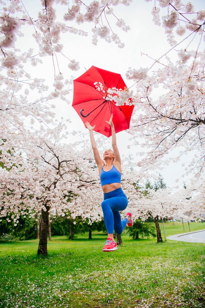 Woman jumping joyfully while reaching for a red umbrella under flowering cherry trees in a grassy park, USA.