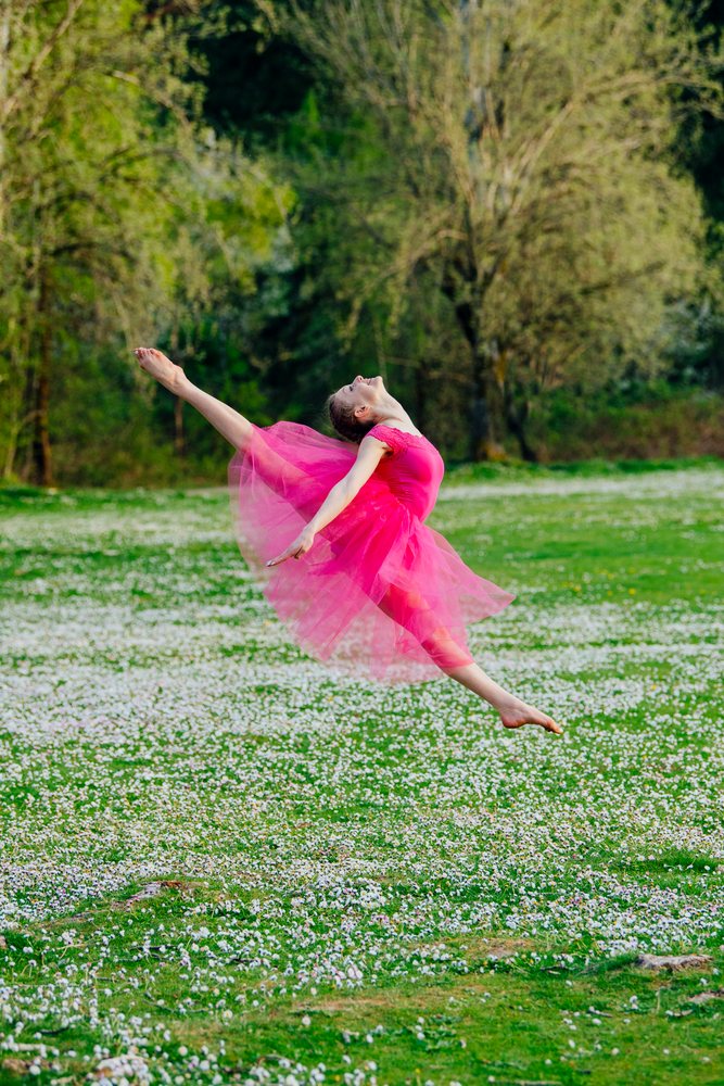 Female ballet dancer wearing a pink tulle dress leaps gracefully barefoot in a green grassy field covered with small white flowers during daytime, USA.