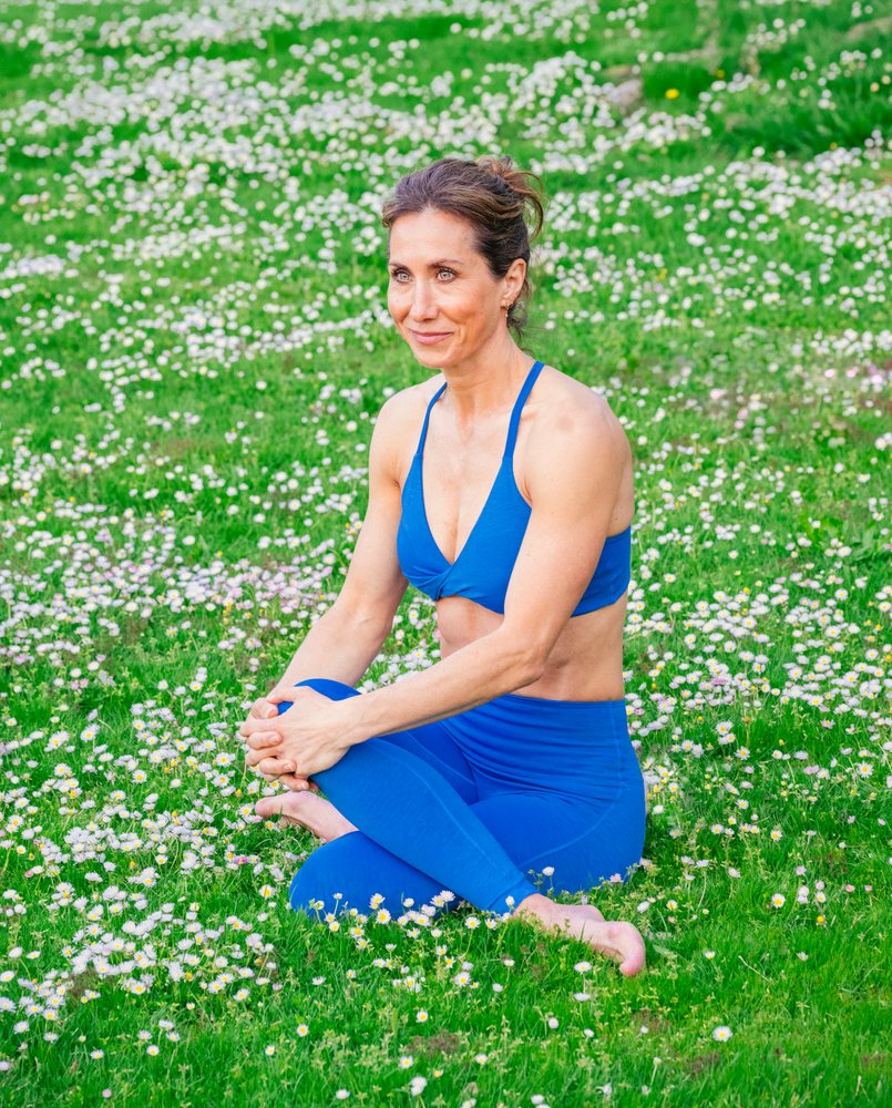 Woman practicing yoga in a seated stretching pose outdoors on a green grassy field with white flowers, USA.