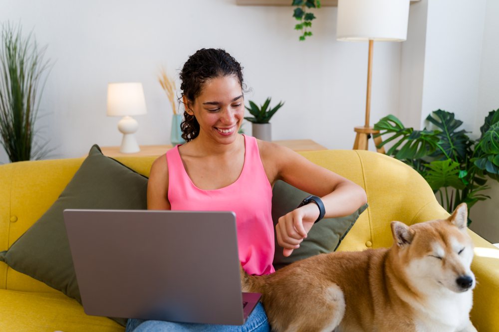 A woman wearing a pink top smiles while sitting on a yellow couch with a laptop and watching her smartwatch next to a medium-sized dog in a living room, Spain.
