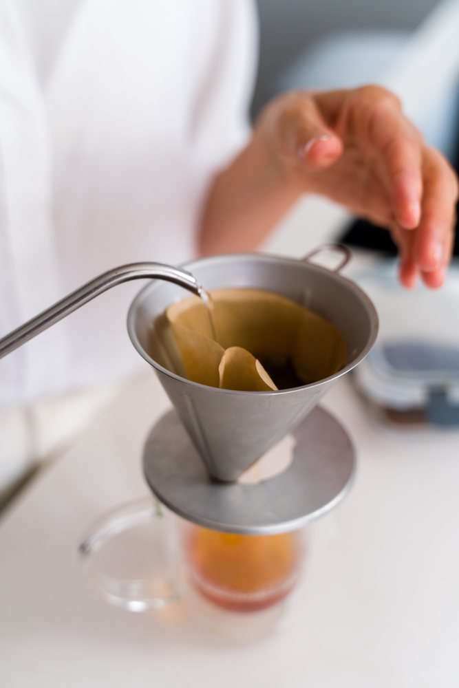 Pouring hot water into a paper coffee filter set in a metal dripper on a glass mug for drip coffee brewing, Spain.