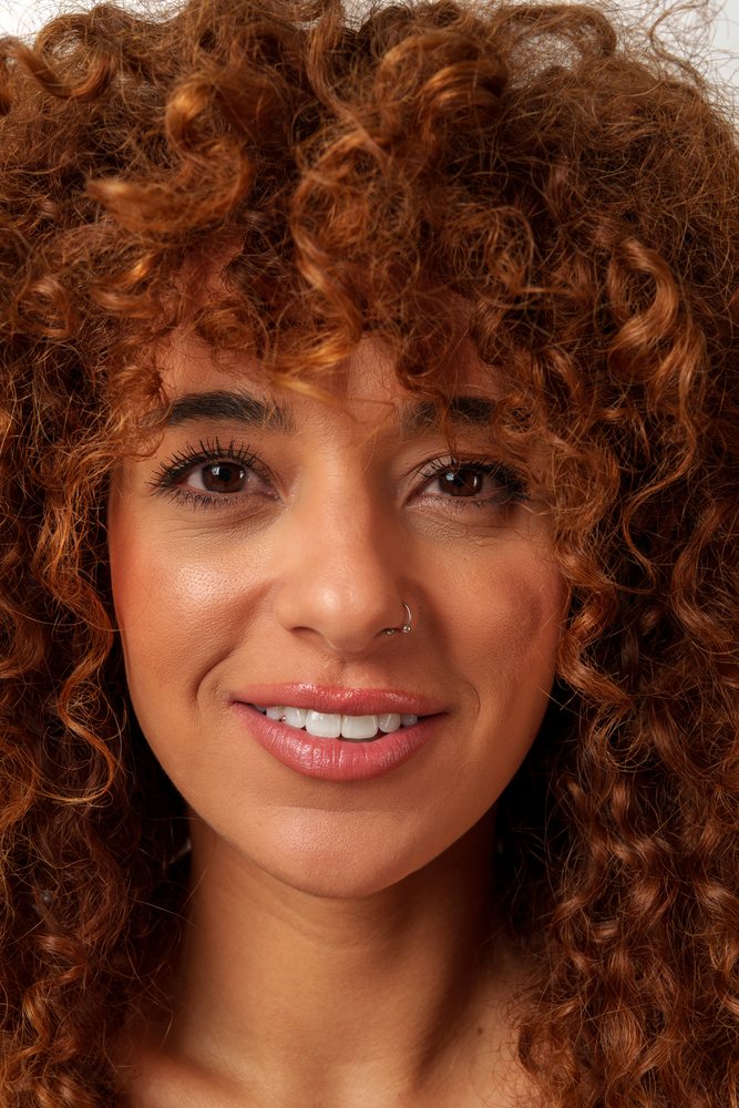 Close-up portrait of a woman with curly brown hair and a nose ring, smiling naturally with visible teeth, Studio, Malaga, Spain.