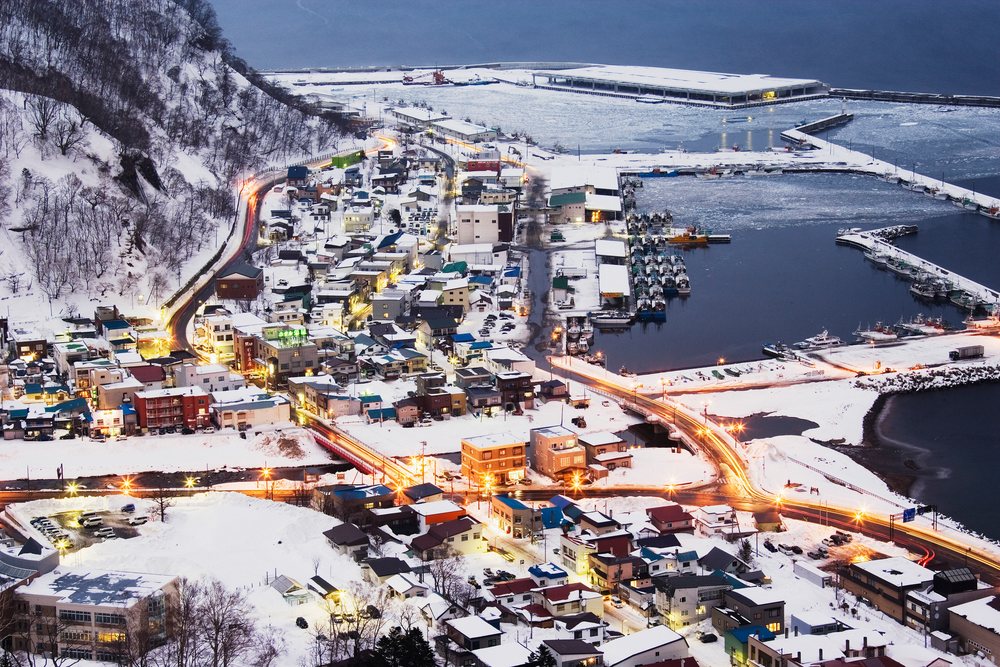 Rausu town skyline in winter shows snowy buildings, harbor with fishing boats, and ocean coastline under evening light, Japan, Asia.